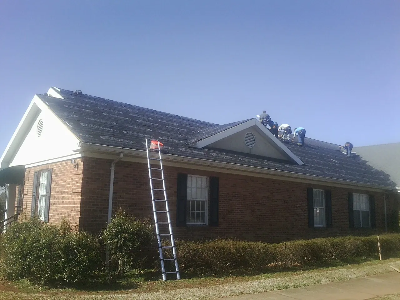 Crew tearing shingles off a brick ranch roof with a ladder set — Emergency Roof Repair work by our crew in Columbus Junction