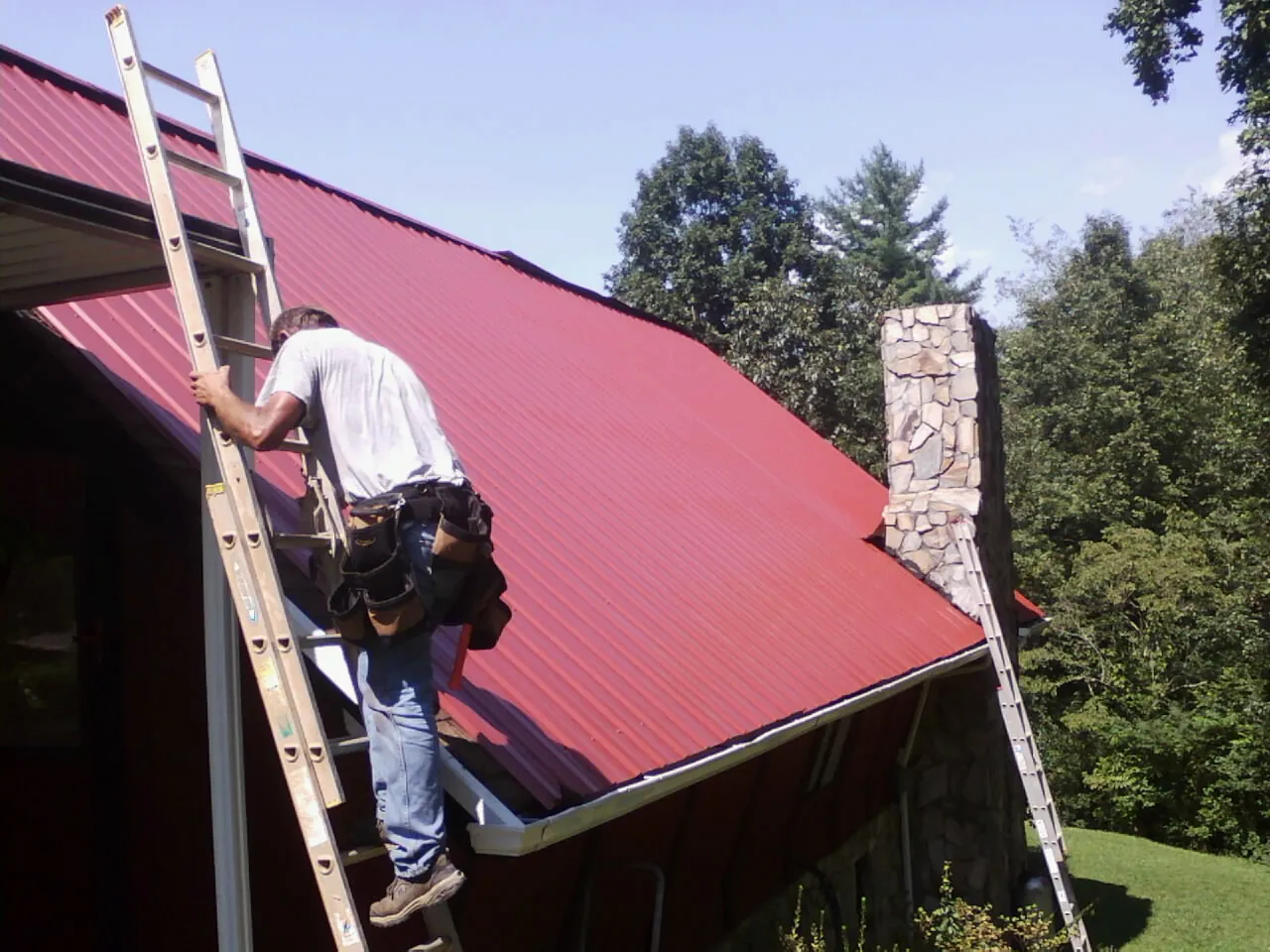 Roofer on a ladder beside a red metal roof and stone chimney — Soffit & Fascia Repair by Roofing Brothers in Columbus Junction